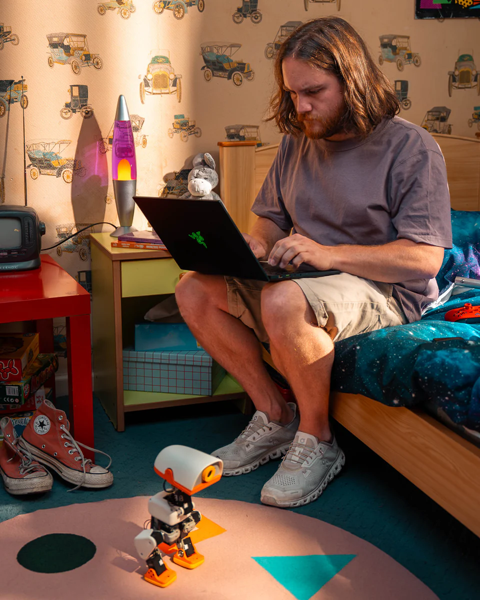 A developer sitting on a bed coding on a laptop while a Microduck stands on the rug in front of them