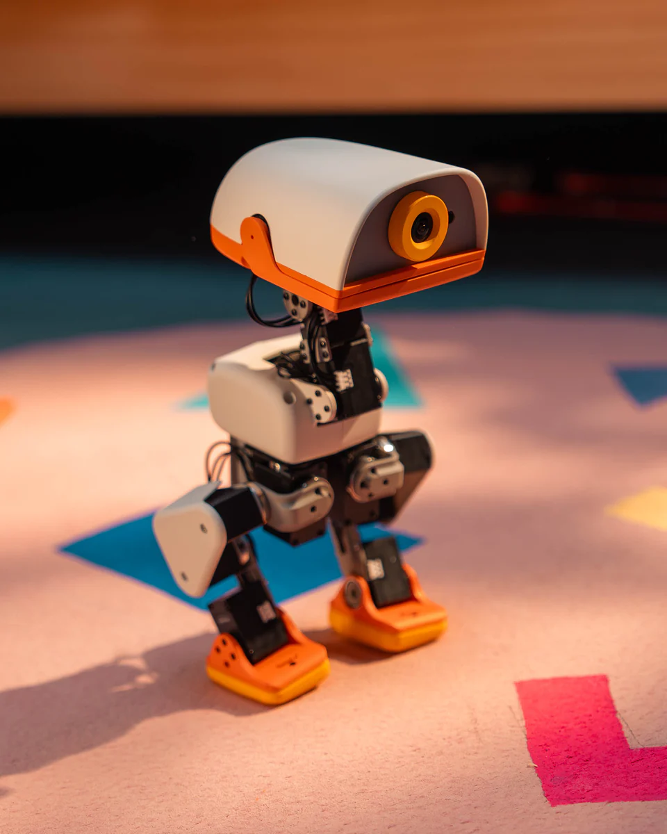 Cream-coloured Microduck robot standing on a patterned playroom rug, its round camera eye and orange beak facing the viewer