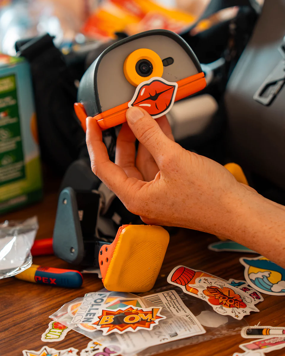 Hands applying a comic-style sticker to a Microduck, with a sheet of stickers spread on the table